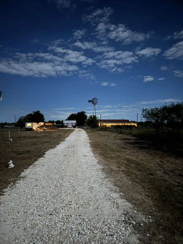 A gravel path leads to a lit farmhouse and windmill under a starry night sky. This is the view coming in from the gate with the boondocking sites on the left. See one visitor already set up.