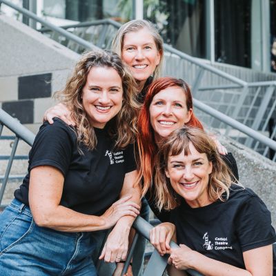 Four smiling women in black t-shirts posing together on stairs.