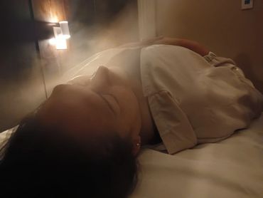 Person lying down in a softly lit room with mist or smoke around.