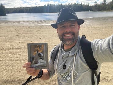 man with gray sweatshirt and wearing fedora holding up a picture of his dog while standing on beach