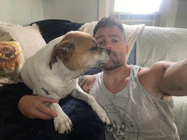 Candid selfie of a man on a couch being kissed by his tan and white dog in a cozy home setting