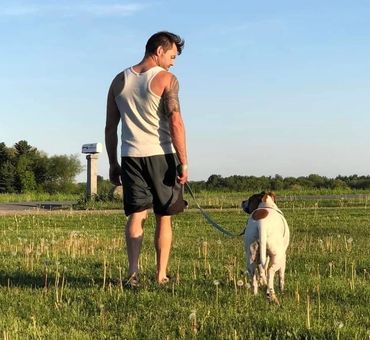 man with white tank top and shorts walking away from camera in grass with a dog by his side