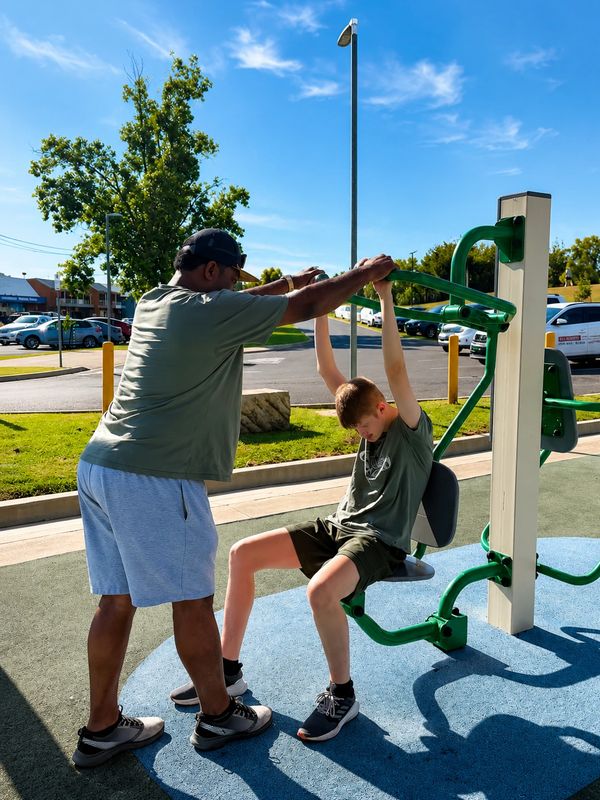 Man helping boy exercise on outdoor gym equipment.