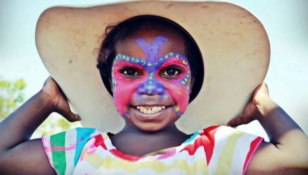 Child with colorful face paint and large hat smiling.