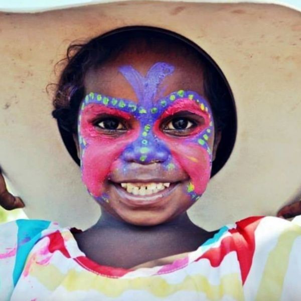 Child with colorful face paint and large hat smiling.