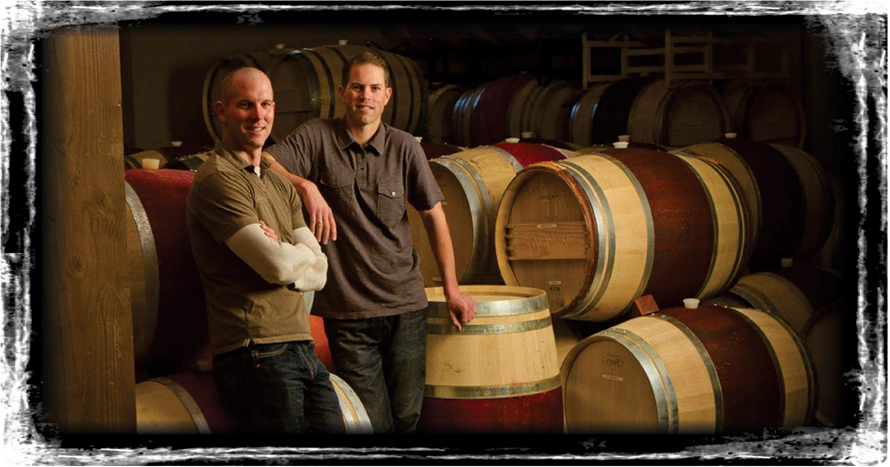 Two winemakers in a cellar full of barrels. 