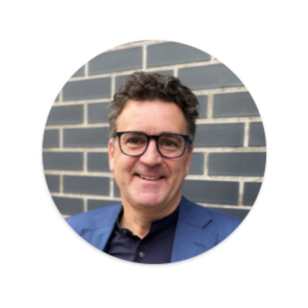 Smiling man in glasses and blue blazer against a brick wall.