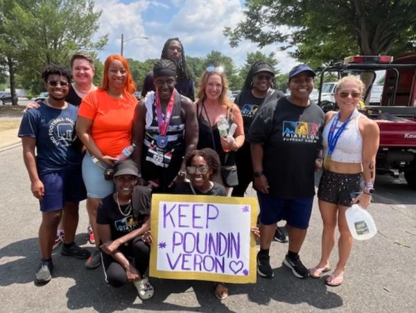 Group of diverse people smiling outdoors, one holding a 'KEEP POUNDIN' sign.