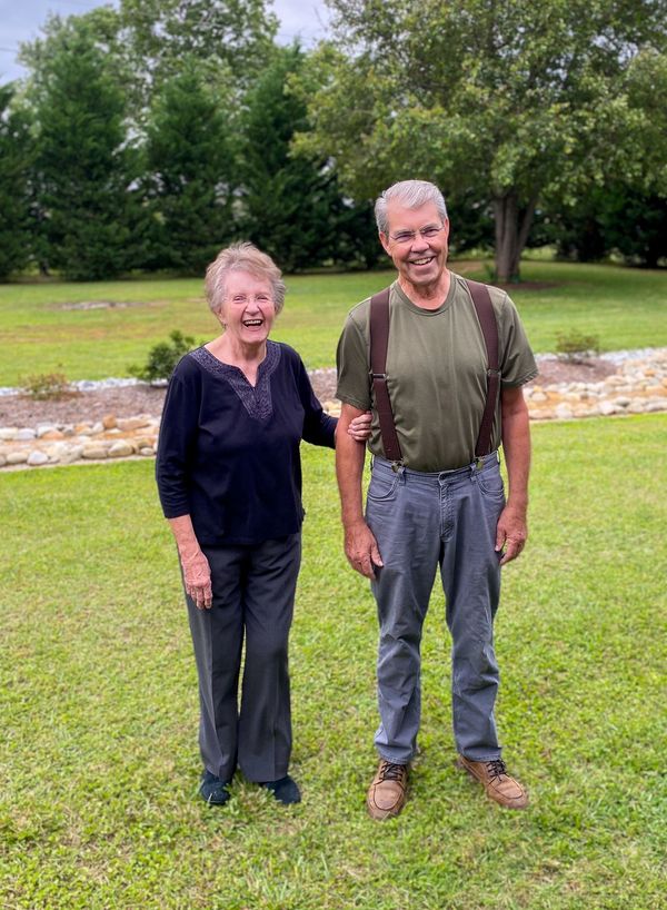 An elderly couple smiling and standing together in a grassy backyard. Remote Controlled Microphone