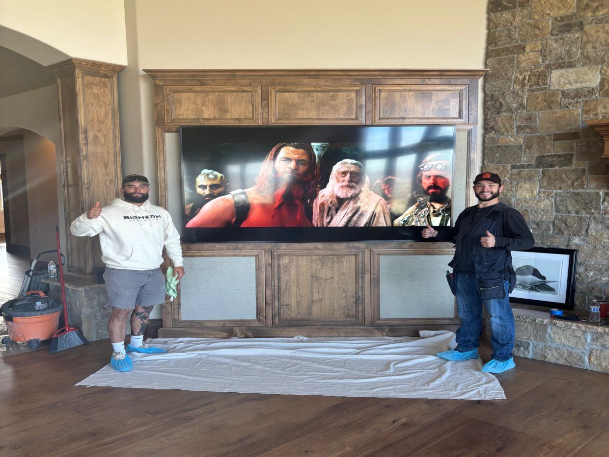 Two men giving thumbs up next to a mounted large TV in a wooden frame.
