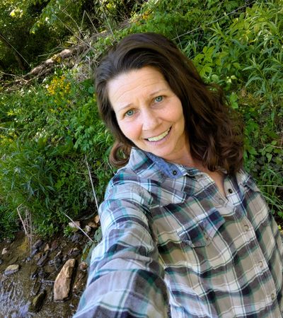 Woman smiling outdoors in a plaid shirt, surrounded by greenery.