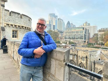 Man in blue jacket smiling by historic stone wall with city skyline in background.