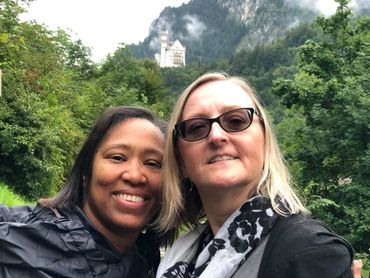 Two women smiling for a selfie with a castle in the misty forested mountains behind them.