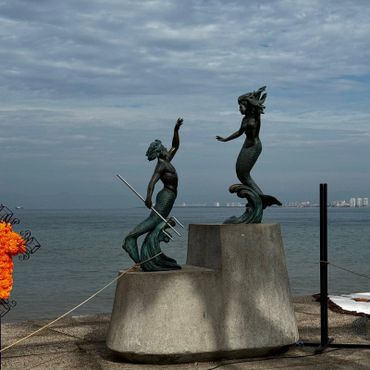 Bronze mermaid and merman statues by the sea on concrete pedestals.