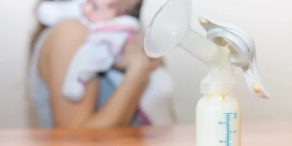 focused picture of manual breast pump with milk. Mother hold baby in background