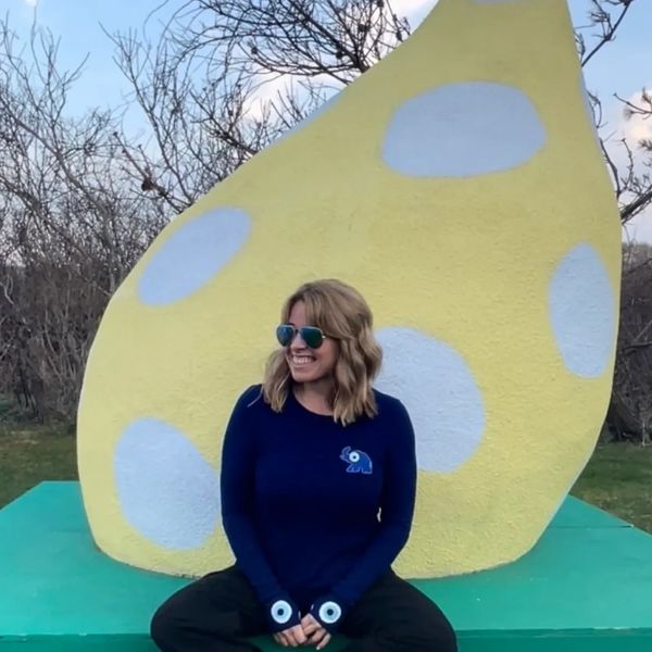Kelly Mitchell smiling and sitting on an art sculpture with trees behind her