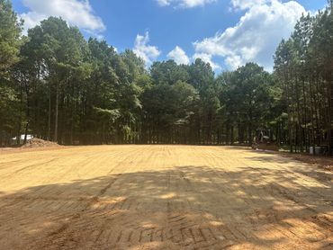Cleared dirt field bordered by trees under a partly cloudy sky.