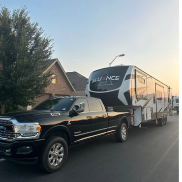 Black pickup truck towing a large Alliance RV trailer in a suburban neighborhood.