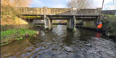 Person in orange jacket measuring water under an old stone bridge over a river.