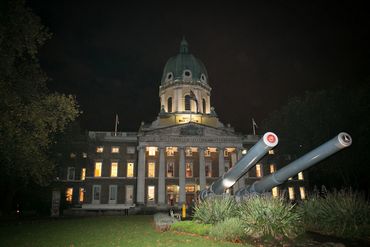 Imperial War Museum building illuminated at night with large cannons in the foreground.