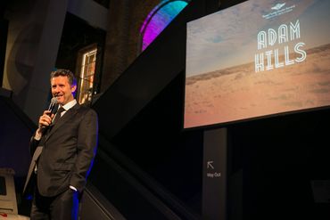 Man in suit speaking into microphone on stage with 'Adam Hills' projected behind him.