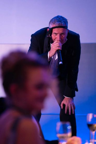 Man in suit singing into microphone with headband, blurred audience in foreground.