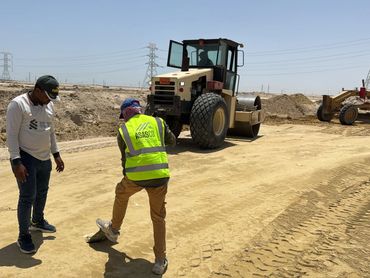 Construction workers and heavy machinery at a sandy site under clear skies.