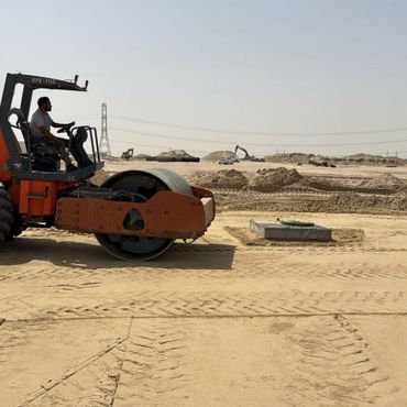 Construction worker operating an orange steamroller on a sandy site.