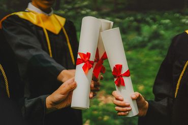 Graduates holding diplomas tied with red ribbons outdoors.