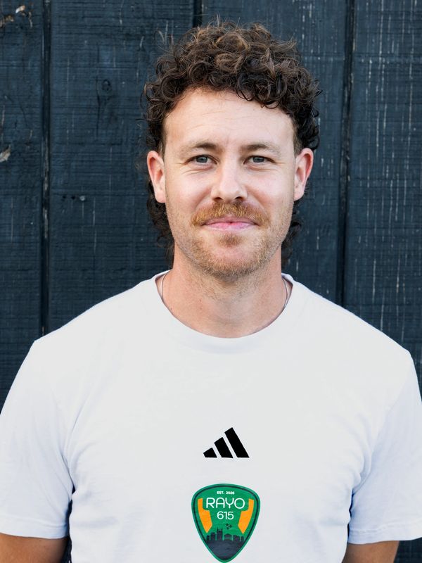Man with curly hair in a white Rayo 615 t-shirt stands against a black wooden wall.