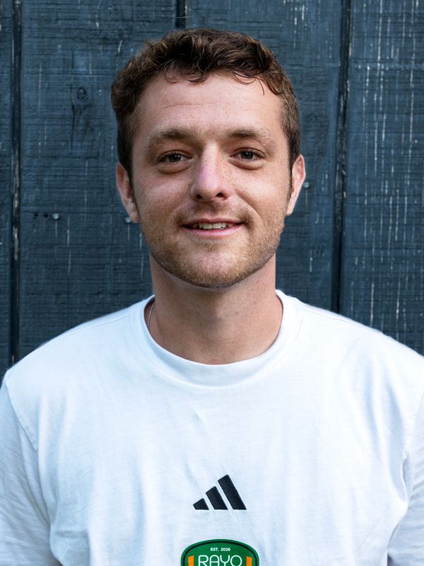 Young man smiling in a white Adidas shirt against a dark wooden background.