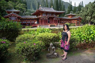 A smiling woman in a floral dress stands near a statue with a traditional Japanese temple in the background.