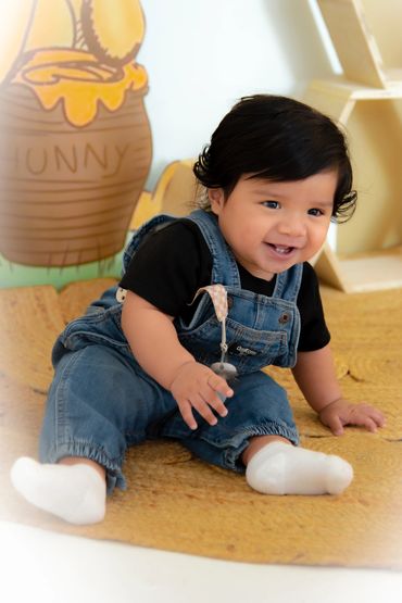 Smiling baby in denim overalls sitting on a yellow mat with a honey pot illustration in the background.