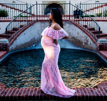 Pregnant woman in a flowing pink dress poses by a decorative water feature.