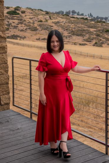 A woman in a red dress stands on a balcony with a dry, hilly landscape in the background.