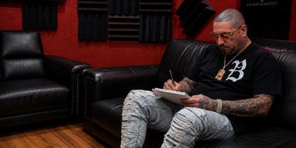 Man with tattoos writing in a notebook on a black couch.