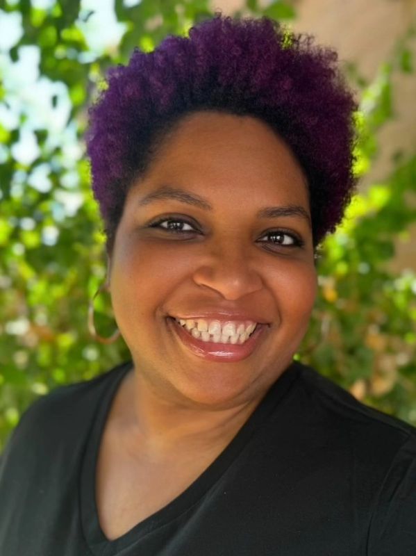 Smiling woman with purple hair and hoop earrings, outdoors with greenery in the background.