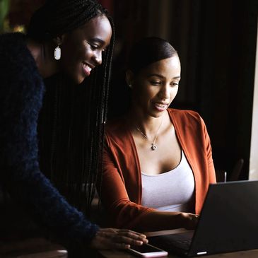 Two woman are looking at a laptop. One is sitting and the other is standing over her.