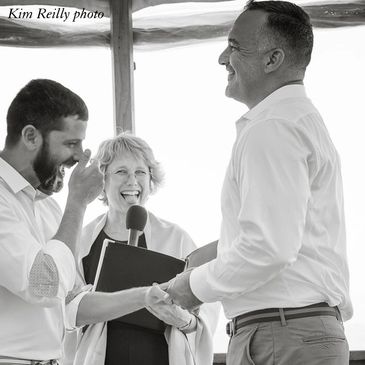 A photograph of two grooms exchanging vows and laughing, with an officiant.