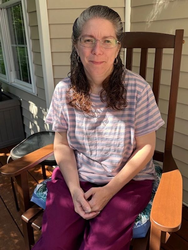 Woman with glasses sitting on a wooden chair outside on a sunny day.