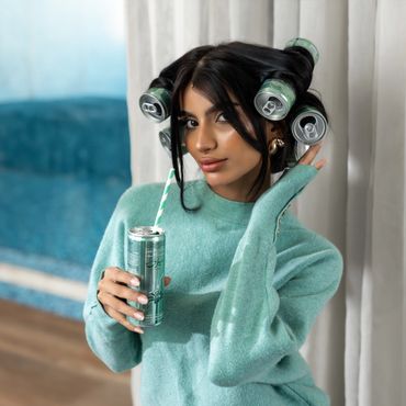 Woman with soda cans in her hair holding a can with a straw, wearing a green sweater.