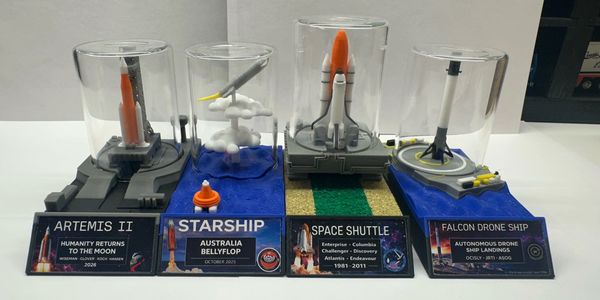 Models of Artemis II, Starship, Space Shuttle, and Falcon Drone Ship under glass cases.