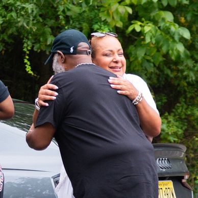 Two people warmly hugging outdoors near cars and greenery.