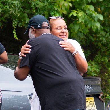 Two people warmly hugging outdoors near cars and greenery.