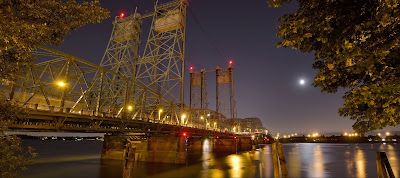 Night view of a lit bridge over water with the moon in the sky.