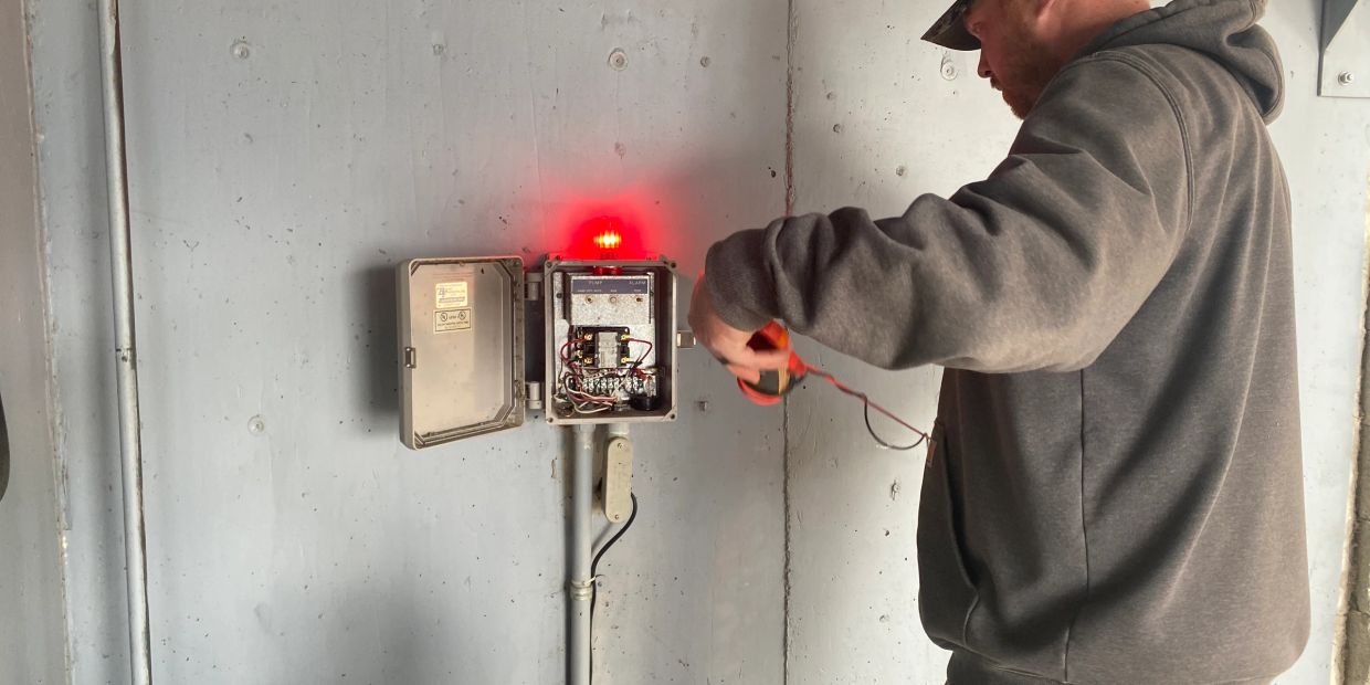 A man works on an electrical control panel with a red warning light.