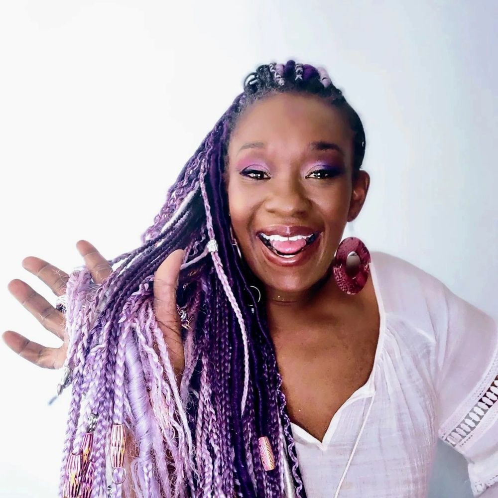 Joyful woman with vibrant purple braids and bright makeup smiles widely.
