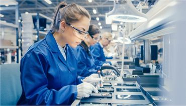 Women assembling smartphones on a factory production line wearing safety glasses and gloves.