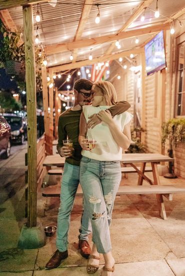 Couple sharing a tender moment outdoors with drinks under warm string lights.