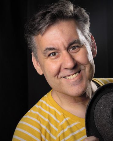 A joyful man in a yellow striped shirt smiling and holding a black hat.